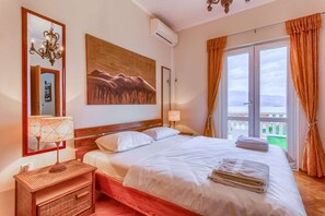 A hotel room with a large bed, a nightstand, a wall-mounted air conditioner, a painting, and a view of the outdoors through a window.