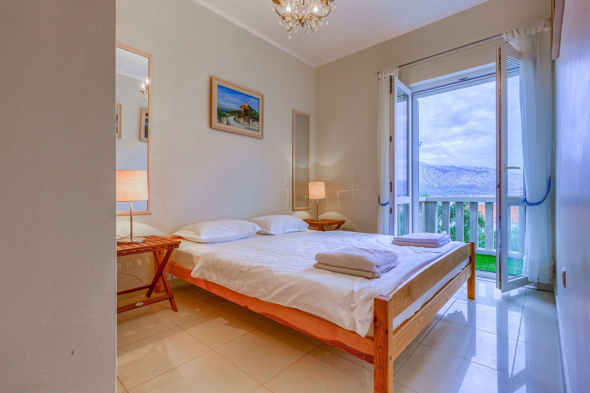 A bedroom with a wooden bed, two bedside tables, a nightstand, a chandelier, a large window with curtains, and a painting on the wall.