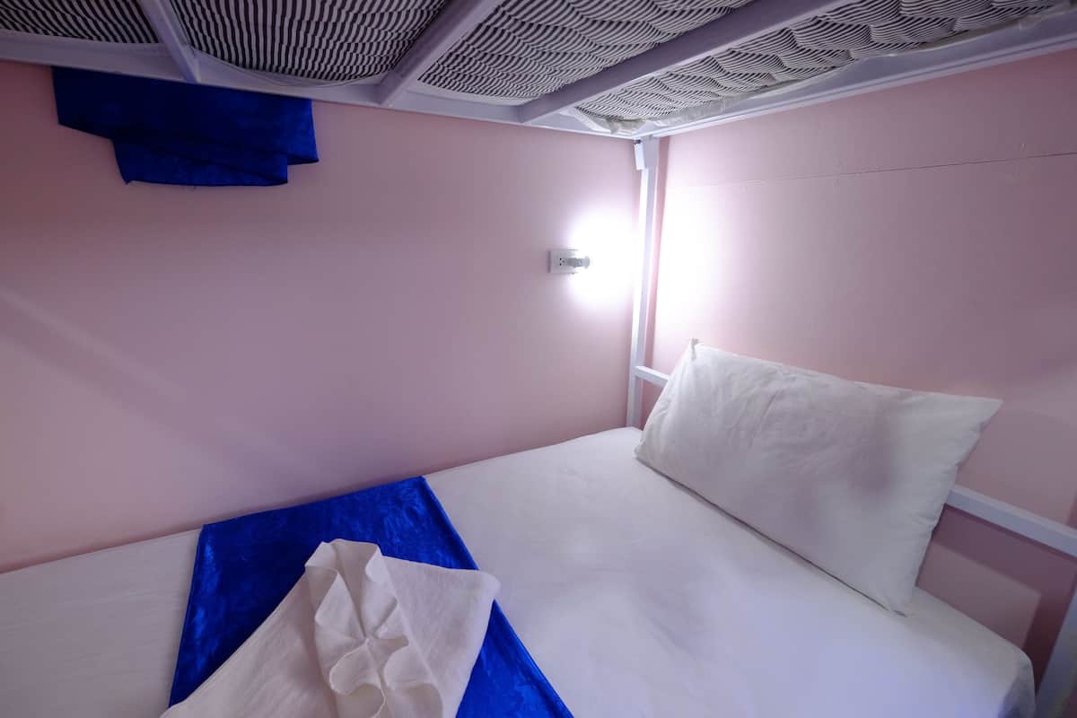 A single bed with white bedding and a blue blanket, a white pillow, and a window with a view of the outside.