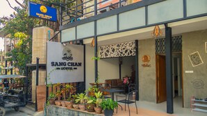 A building with a sign reading "SANG CHAN HOSTEL" and a welcome sign. There are potted plants in front, a table and chairs outside, and a staircase leading to an upper level. - Sang Chan Hostel (Koh Lipe)
