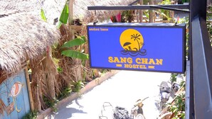 A sign for Sang Chan Hostel with a thatched-roof structure in the background. - Sang Chan Hostel (Koh Lipe)