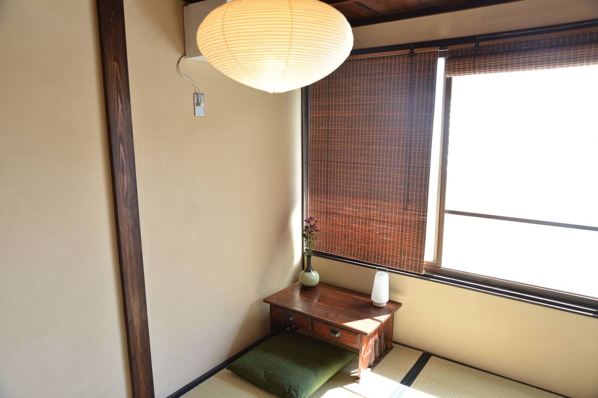 A traditional Japanese room with tatami flooring, a shoji screen, a wooden desk with a vase and a lamp, and a hanging paper lantern.