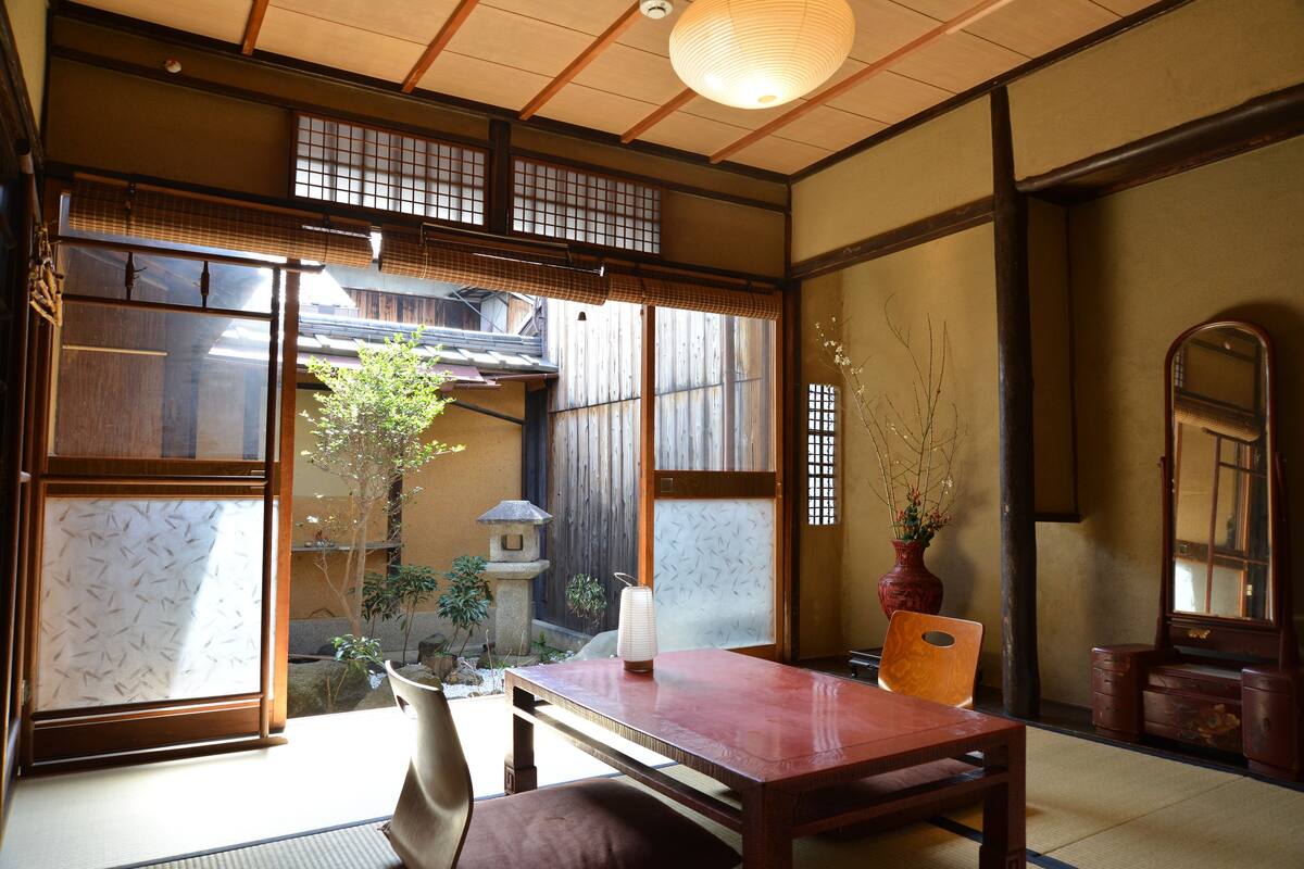 A traditional Japanese room with tatami flooring, a wooden table, a chair, a vase with branches, and a view of a garden through shoji screens.