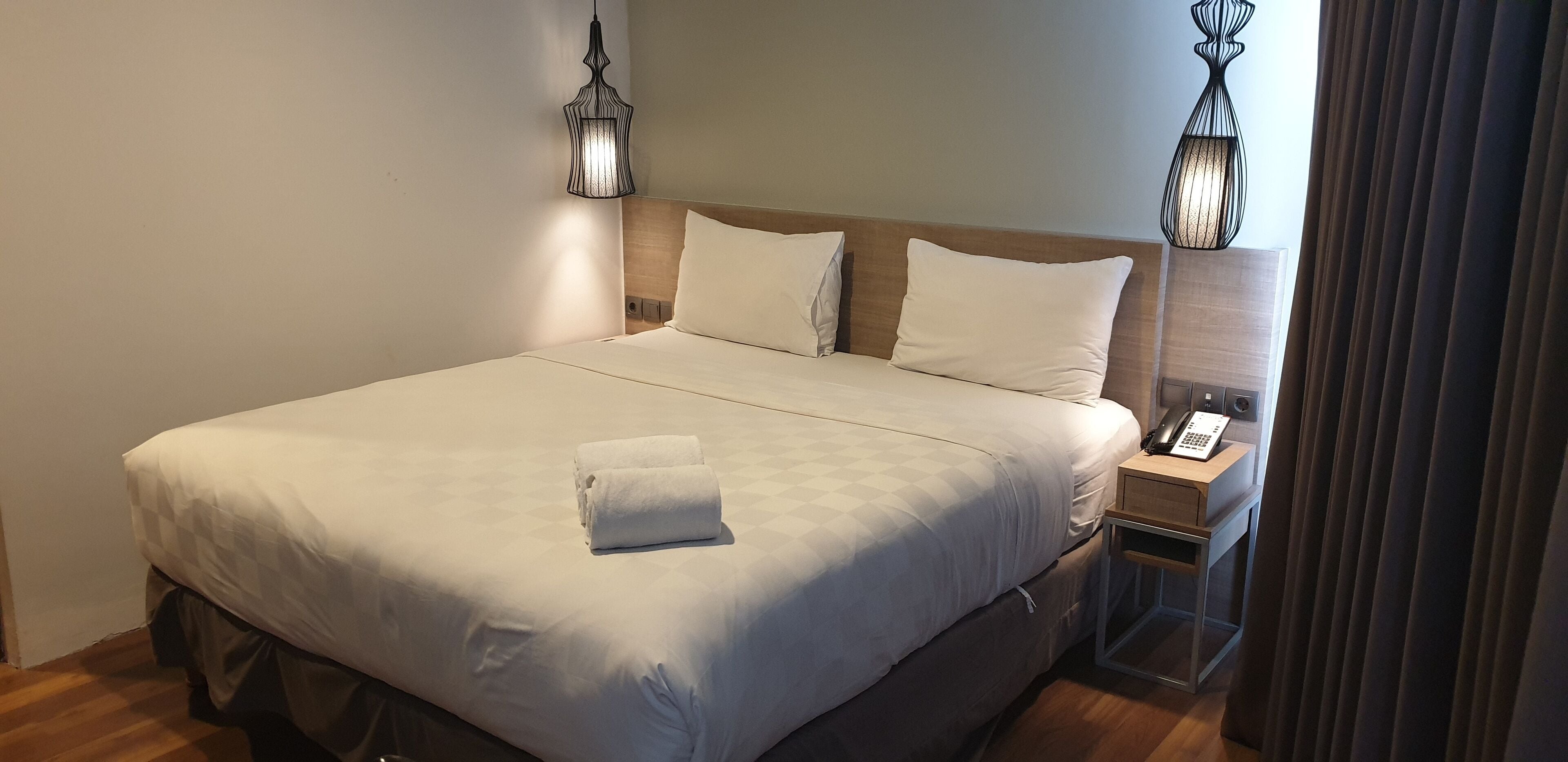 Un lit bien fait, avec du linge de lit blanc et une serviette pliée, une table de chevet avec un téléphone et une lampe, et une tête de lit en bois.