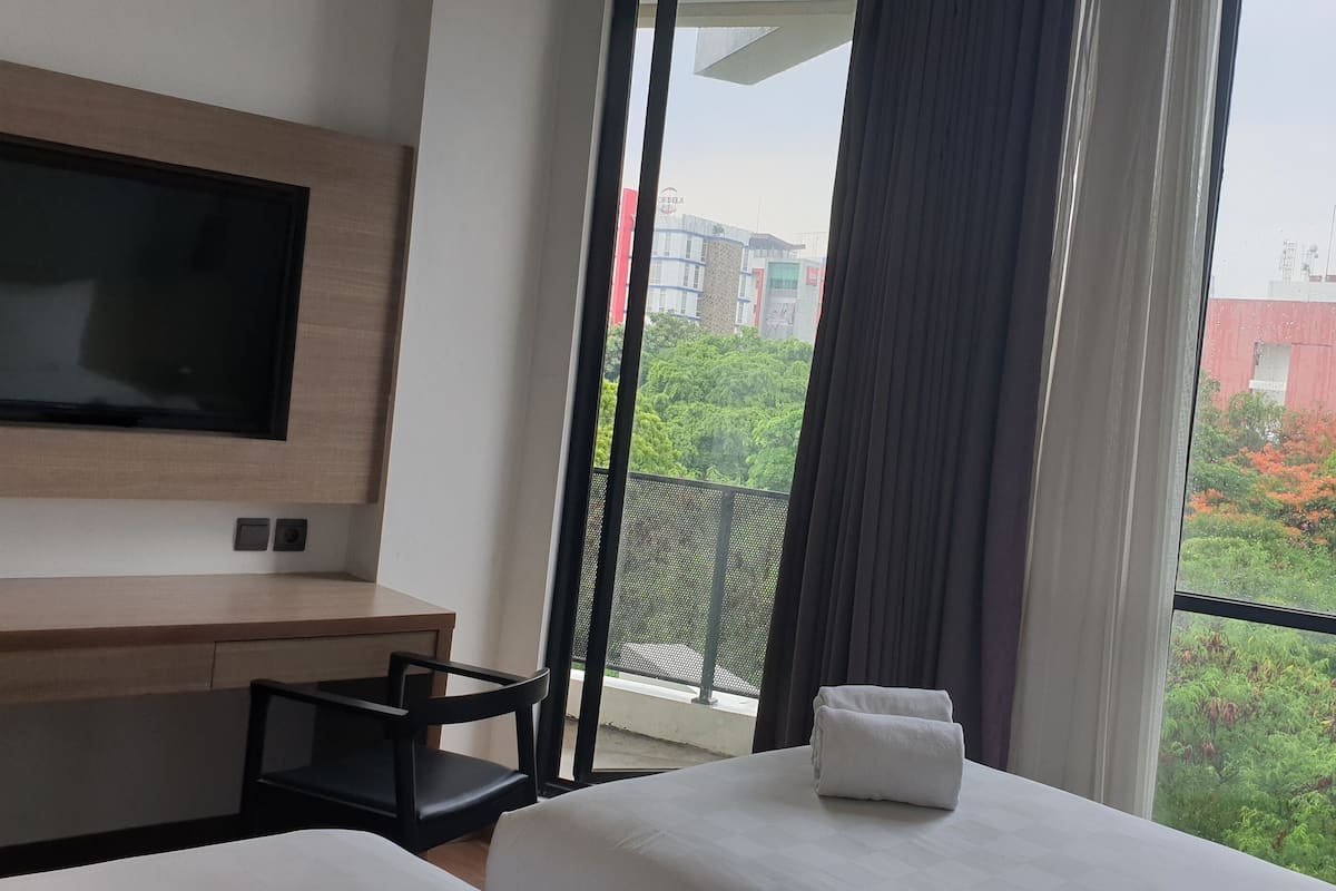 A hotel room with a bed, a TV mounted on the wall, and a balcony with a view of buildings and trees.