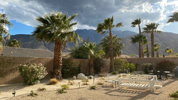 An outdoor patio area with palm trees, a brick wall, and a mountainous backdrop. - Sunny Palm Springs Solar Oasis 180° Mountains, 30ft Palms & Boulder Desertscape (Palm Springs)