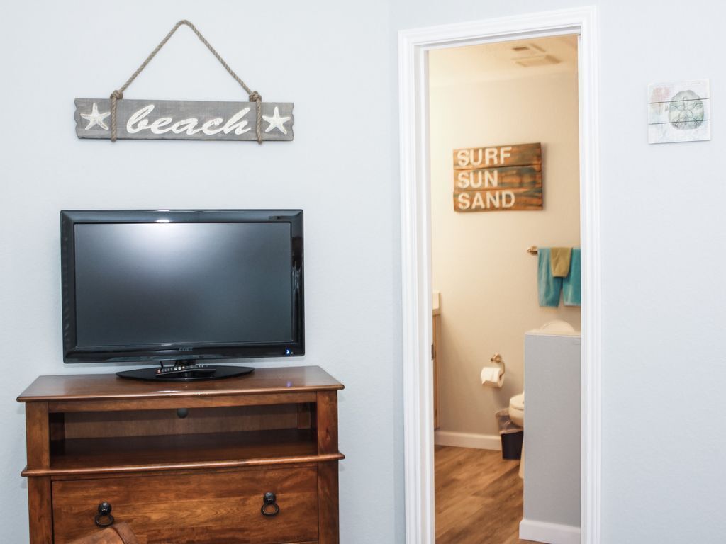 Un televisor sobre un soporte de madera, un letrero con la palabra 'playa' y un letrero que dice 'SURF SUN SAND' en una habitación con una puerta que da a un baño.