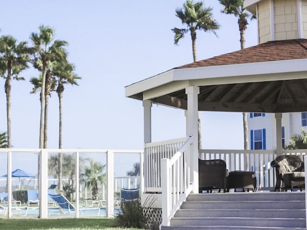 A covered outdoor area with a pool, palm trees, and a building in the background.