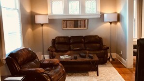 A living room with a leather sofa, a coffee table, and a lamp. There are windows with white frames and a wooden headboard above the sofa.
