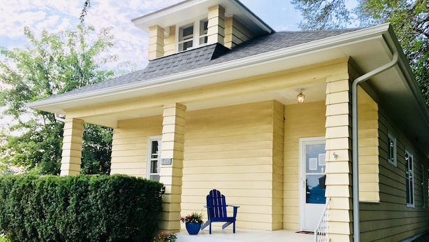 A two-story house with a covered porch, a blue chair, and potted plants.
