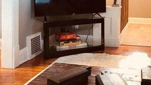 A living room with a flat-screen TV on a stand, a small table with board games, and a staircase leading to another room.
