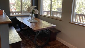 A wooden table with a vase of flowers, two windows with views of a deck and trees, and a bench.