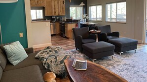 A spacious living area with a sofa, ottoman, and a coffee table. A kitchen with wooden cabinets and modern appliances is visible in the background.