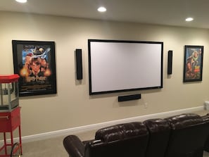Sala de cinema com uma TV de tela grande, dois pôsteres na parede, um carrinho de pipoca vermelho e um sofá de couro marrom.