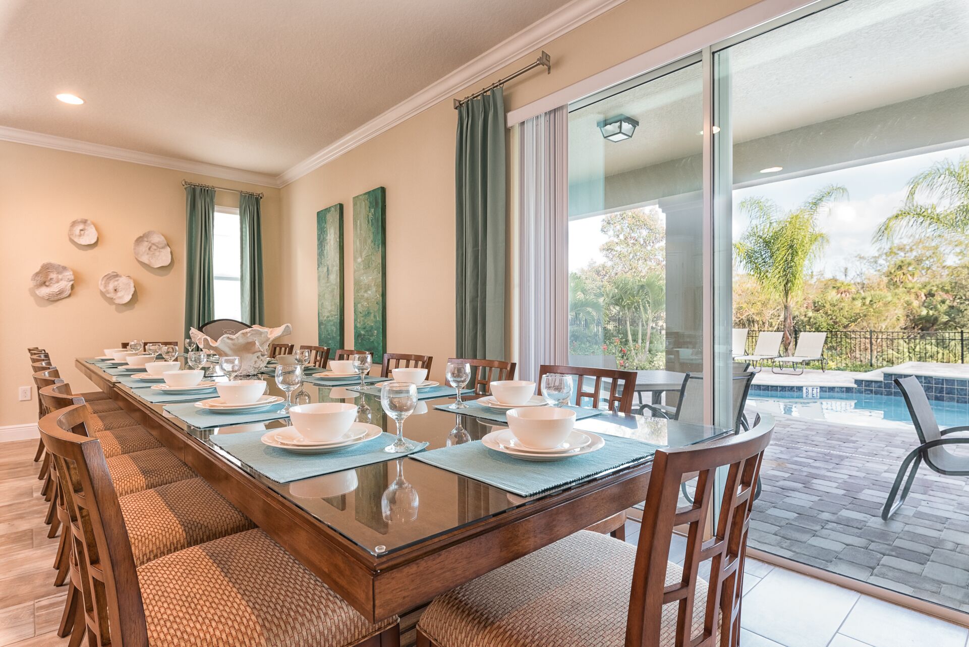 Une salle à manger avec une longue table dressée pour un repas, de grandes fenêtres et une vue sur une piscine.