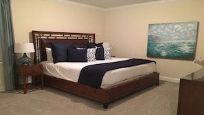 Cama bem arrumada com cabeceira de madeira, mesa de cabeceira com abajur e pintura de paisagem marinha na parede.
