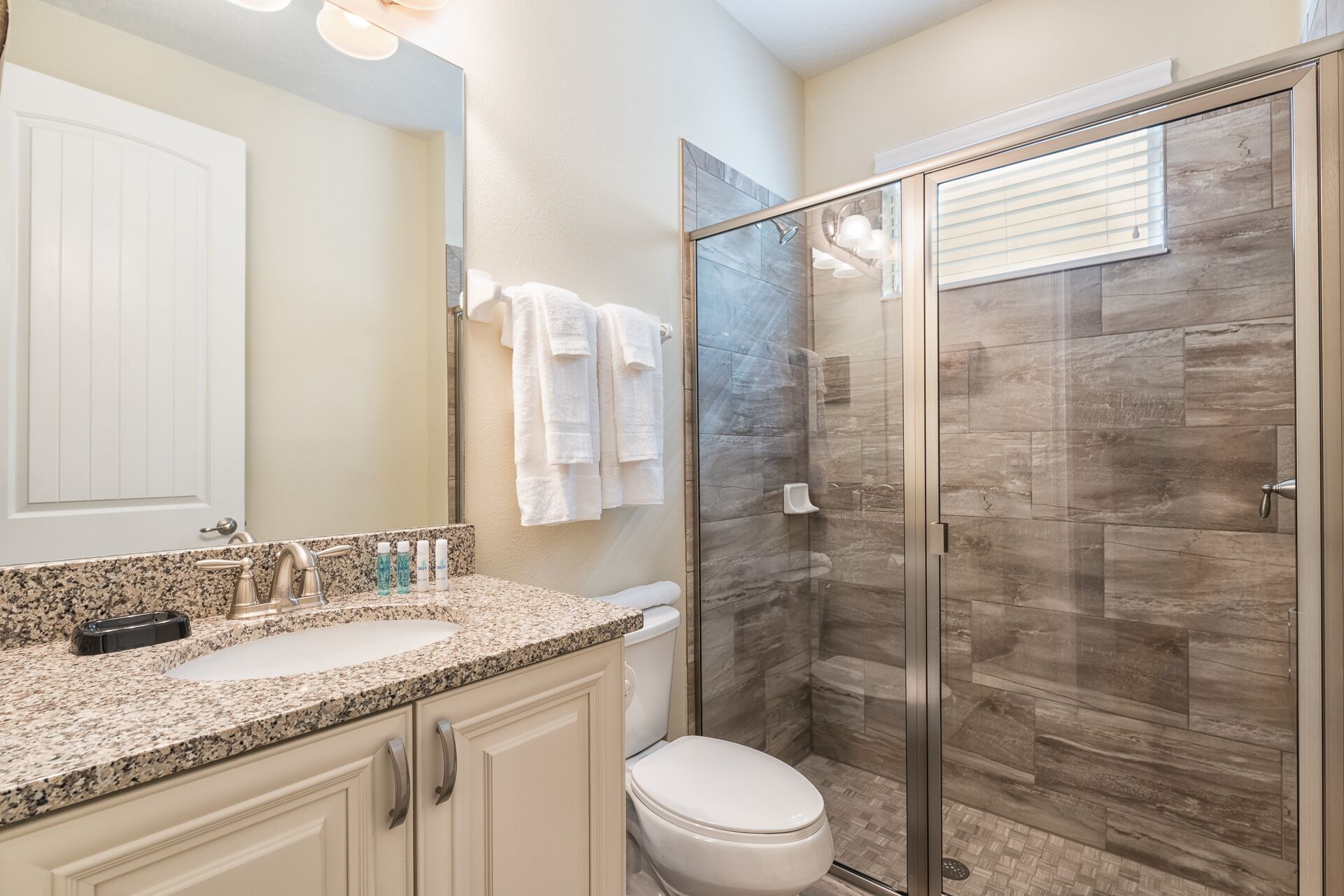 A bathroom with a granite countertop, a toilet, a glass shower enclosure, and a mirror.