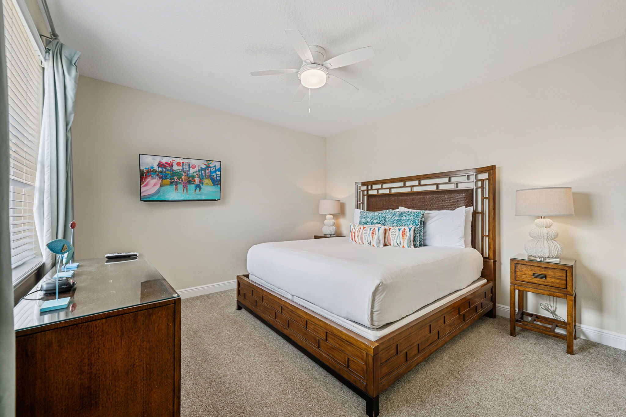 Une chambre à coucher avec un lit en bois, un bureau, une chaise, une table de chevet, une lampe, un ventilateur de plafond et un tableau accroché au mur.