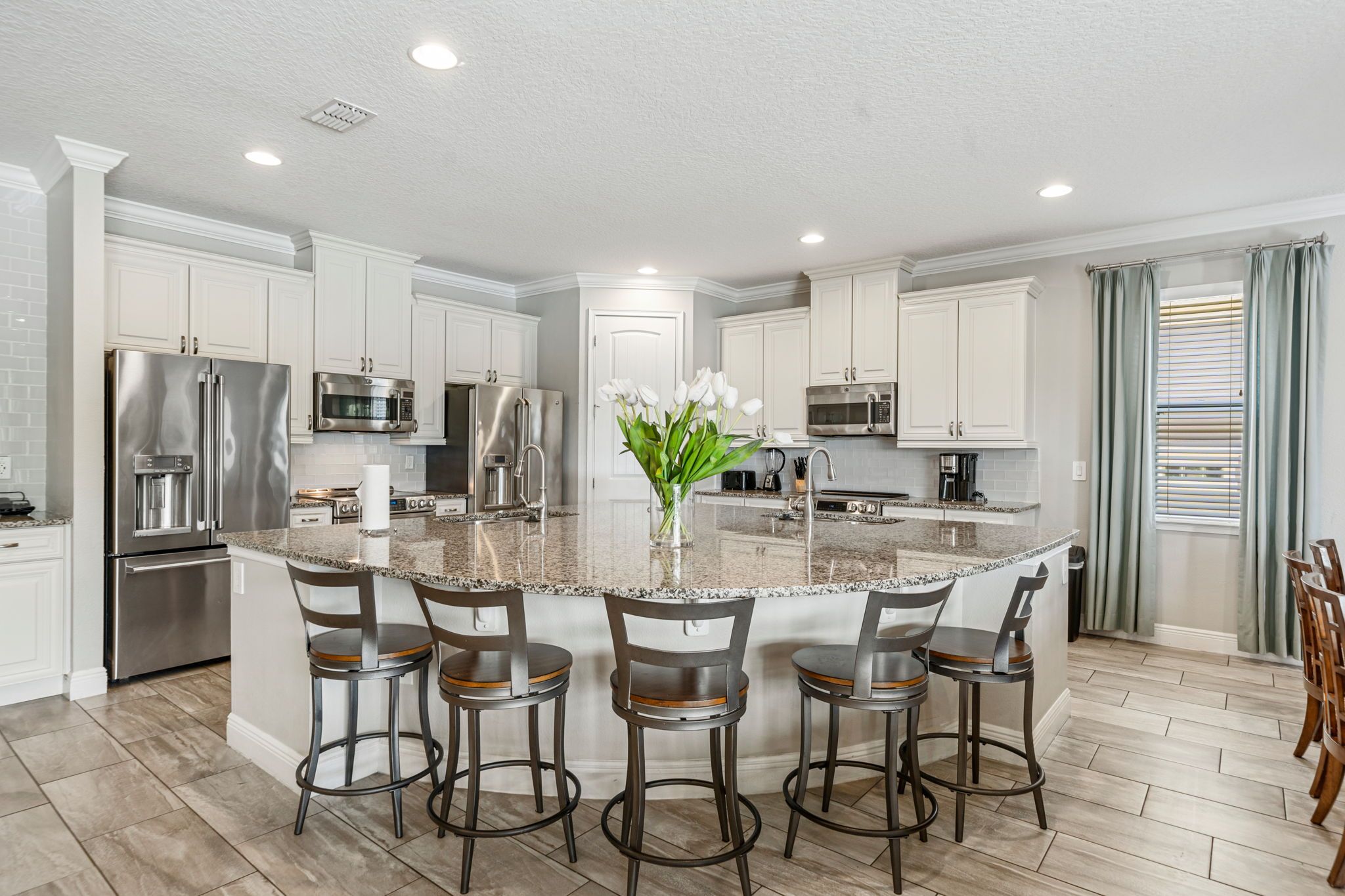 A modern kitchen with white cabinetry, stainless steel appliances, a granite island with bar stools, and a vase of flowers.