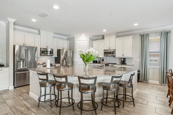 A modern kitchen with white cabinetry, stainless steel appliances, a granite island with bar stools, and a vase of flowers.