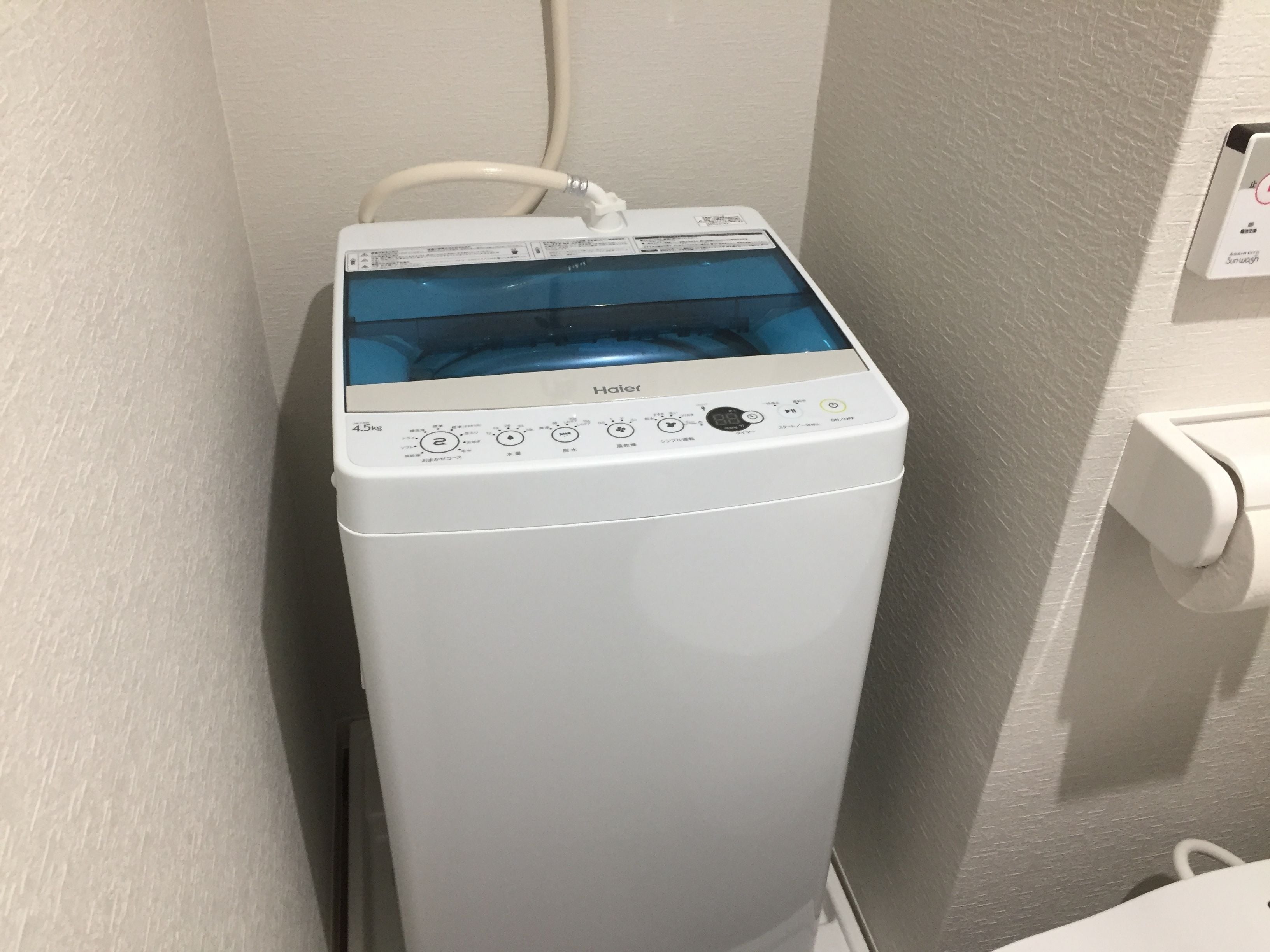 Una lavadora blanca, compacta, de carga frontal, con pantalla digital y panel de control, ubicada en un rincón de una habitación con paredes lisas.