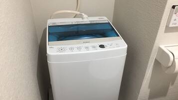 Una lavadora blanca, compacta, de carga frontal, con pantalla digital y panel de control, ubicada en un rincón de una habitación con paredes lisas.