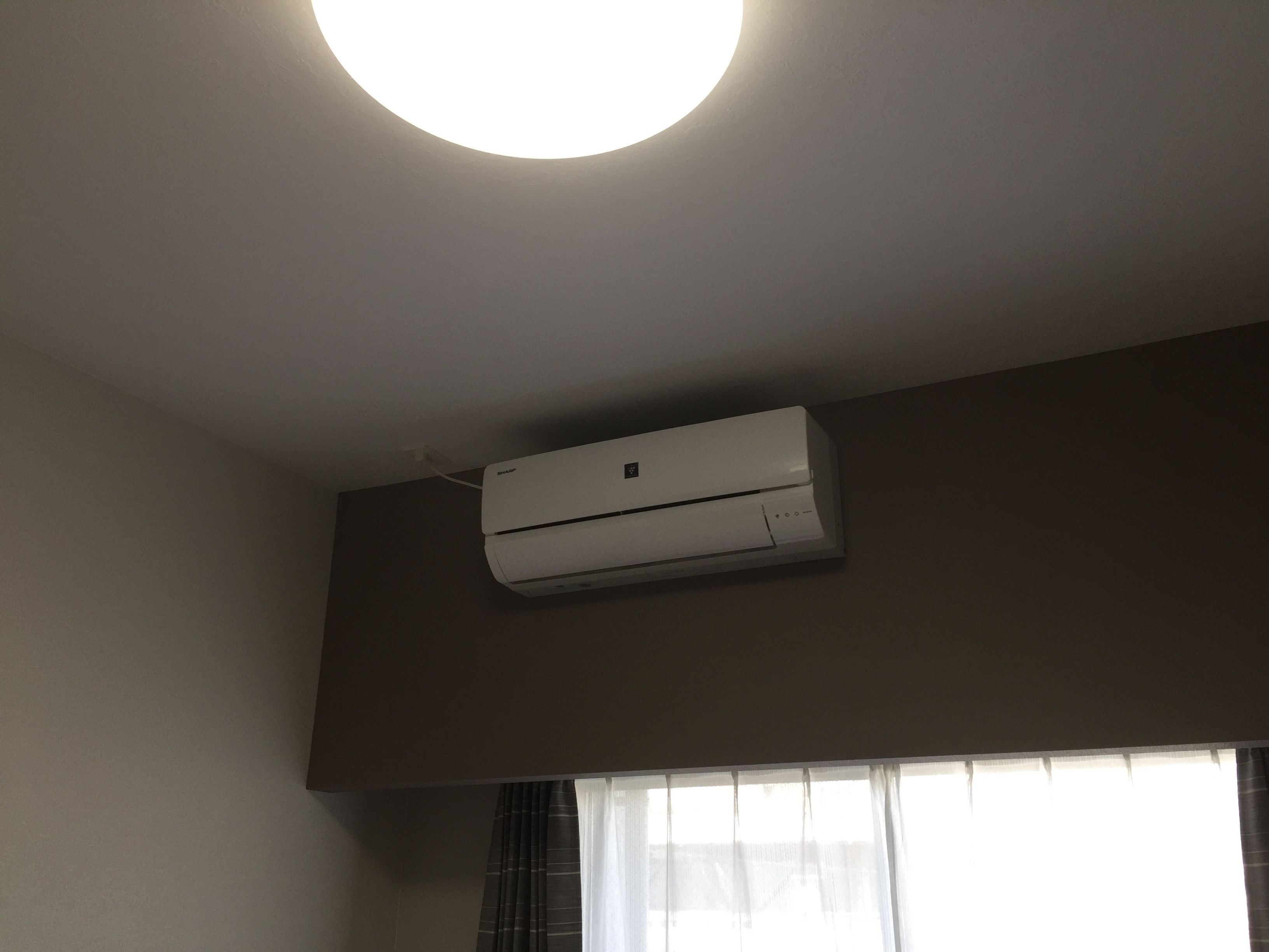 Quarto com ar-condicionado instalado no teto, janela com cortinas leves e parede lisa.