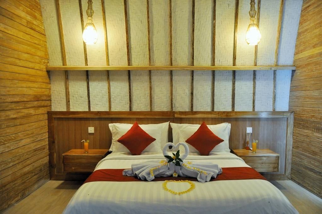 A neatly made bed with red pillows, a heart-shaped decoration, and a towel arrangement. There are two bedside tables with lamps and a wooden headboard.
