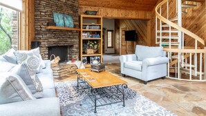 Una sala de estar acogedora con chimenea de piedra, una escalera de madera y una estantería llena de libros y objetos decorativos.