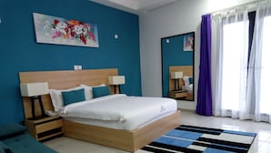 A hotel room with a large bed, two bedside tables with lamps, a blue wall, a floral painting, a mirror, and a window with purple curtains. - Résidence Ethan (Abidjan)
