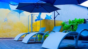 A row of white lounge chairs under a large umbrella, with a yellow wall and a mural in the background. - Résidence Ethan (Abidjan)