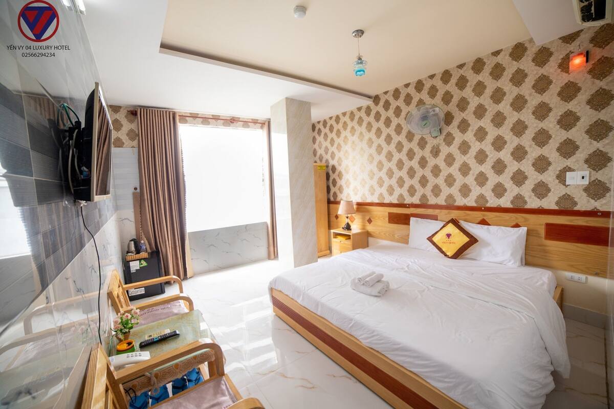 Una habitación de hotel con cama, televisión montada en la pared, mesita de noche con lámpara, cabecero de madera y ventana con cortinas.
