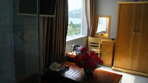 A room with a wooden desk, a telephone, a vase with red flowers, a mirror, a wooden wardrobe, and a window with a view of greenery and mountains. - Yen Vy 04 Luxury Hotel (Quy Nhon Nam)