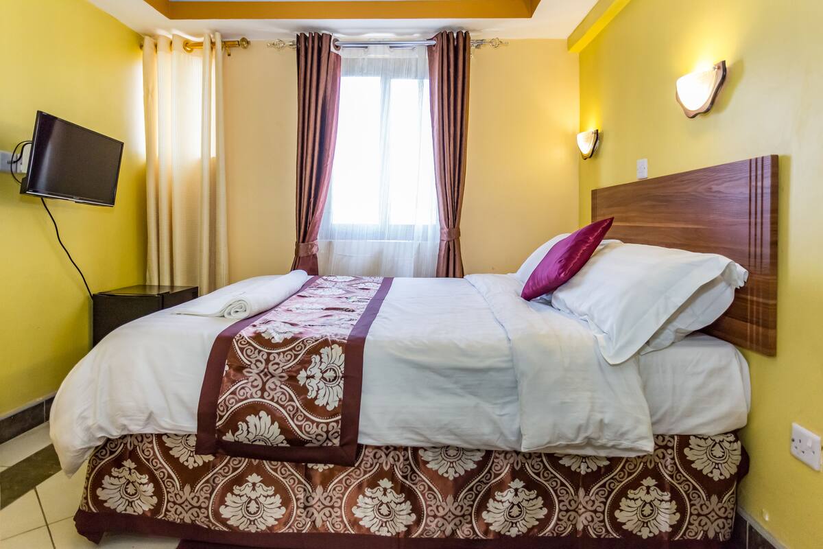 A neatly made bed with a patterned bedspread and pillows, a flat-screen TV mounted on the wall, and a window with curtains.