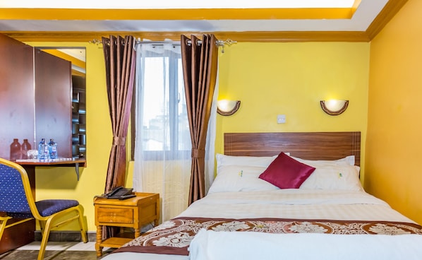 Une chambre d’hôtel comprenant un lit, un bureau avec une chaise, un téléphone et une étagère avec des bouteilles.