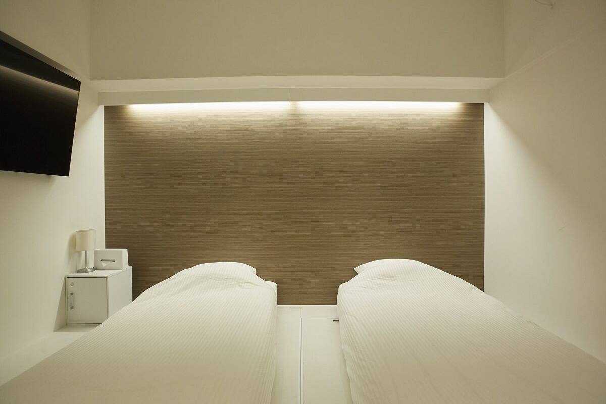 A hotel room with two single beds, a bedside table with a lamp, and a flat-screen TV mounted on the wall.