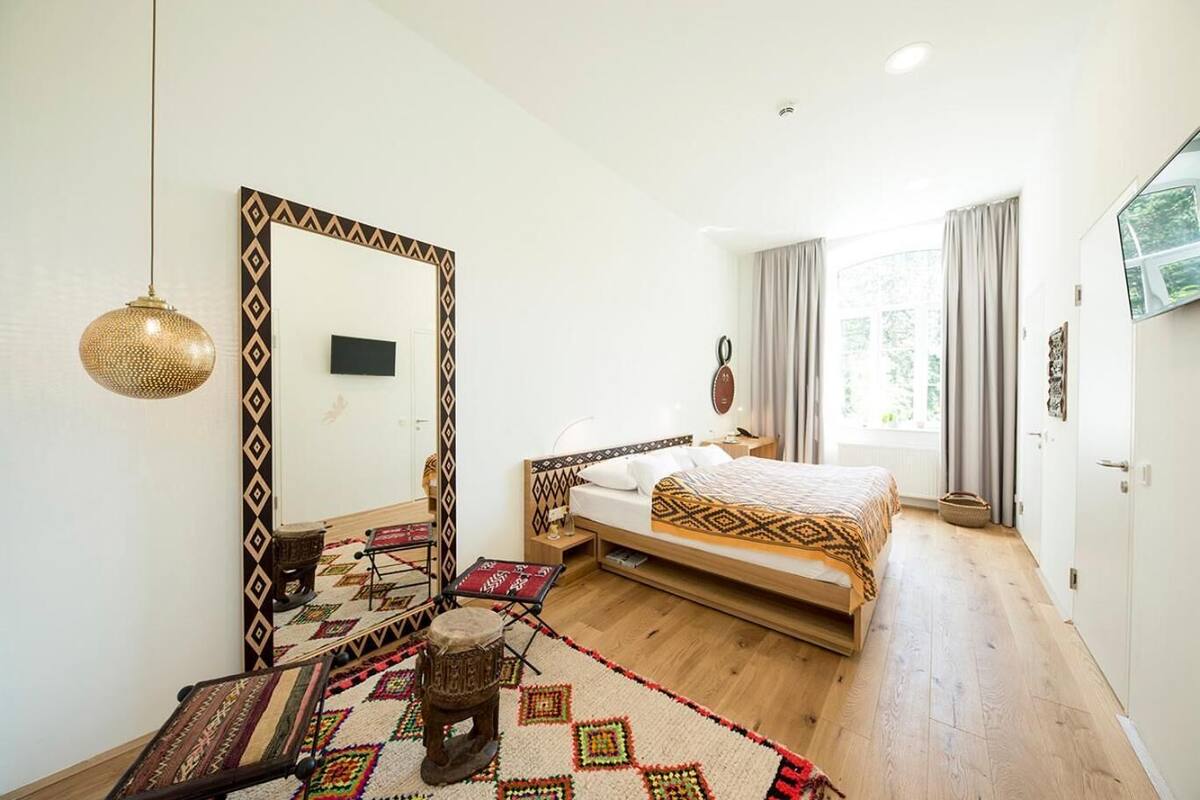 Un dormitorio con un espejo grande, una cama con ropa de cama estampada, piso de madera y vistas al exterior a través de una ventana.