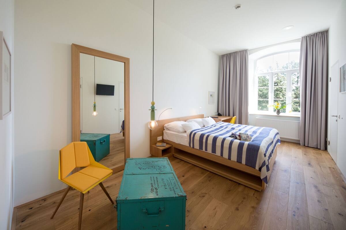 Un dormitorio con cama, una silla, un escritorio, un espejo y una ventana.