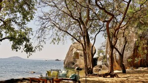 A beach setting with a table and chairs under a tree, a large rock formation, and a view of the sea.
