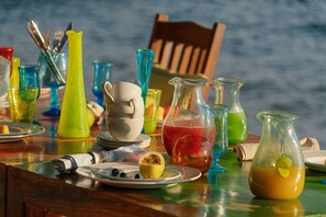 A table set with colorful glassware, plates, and a pitcher of orange juice, with a wooden chair and a view of the sea in the background. - Lukuba Island Lodge - East Africa Camps (Musoma)