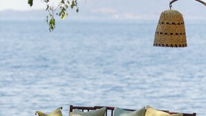 A beachside seating area with a wooden bench and cushions, a woven hanging light fixture, and a view of the sea and mountains in the background.