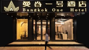L’extérieur du Bangkok One Hotel présente un design moderne, avec une grande enseigne lumineuse, une entrée en verre et un intérieur bien éclairé, visible à travers les portes.