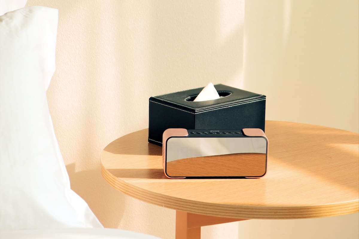 Un lit bien fait, avec du linge de lit blanc, une boîte de mouchoirs noire posée sur une table de chevet en bois, et un mur sur lequel se projette l’ombre d’une fenêtre.