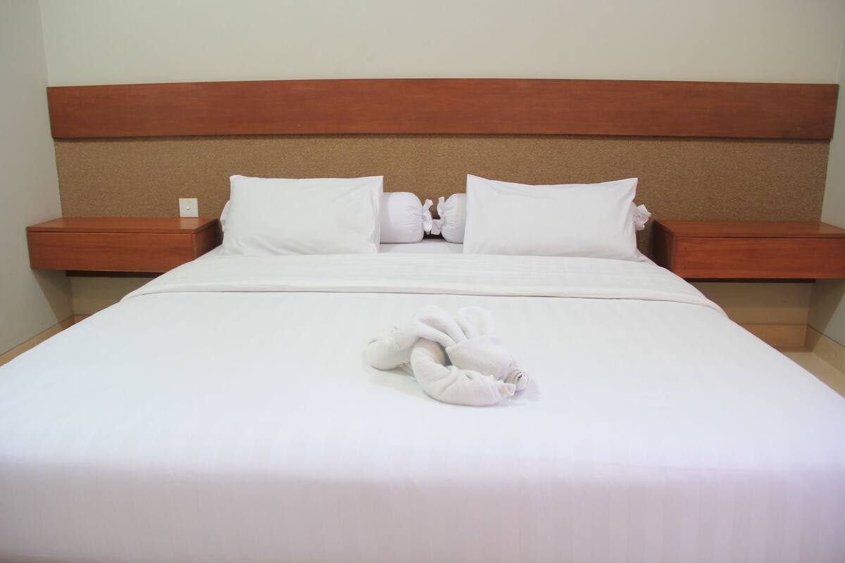 A neatly made bed with white linens and two pillows, a wooden headboard, and two wooden nightstands with a small table on each.