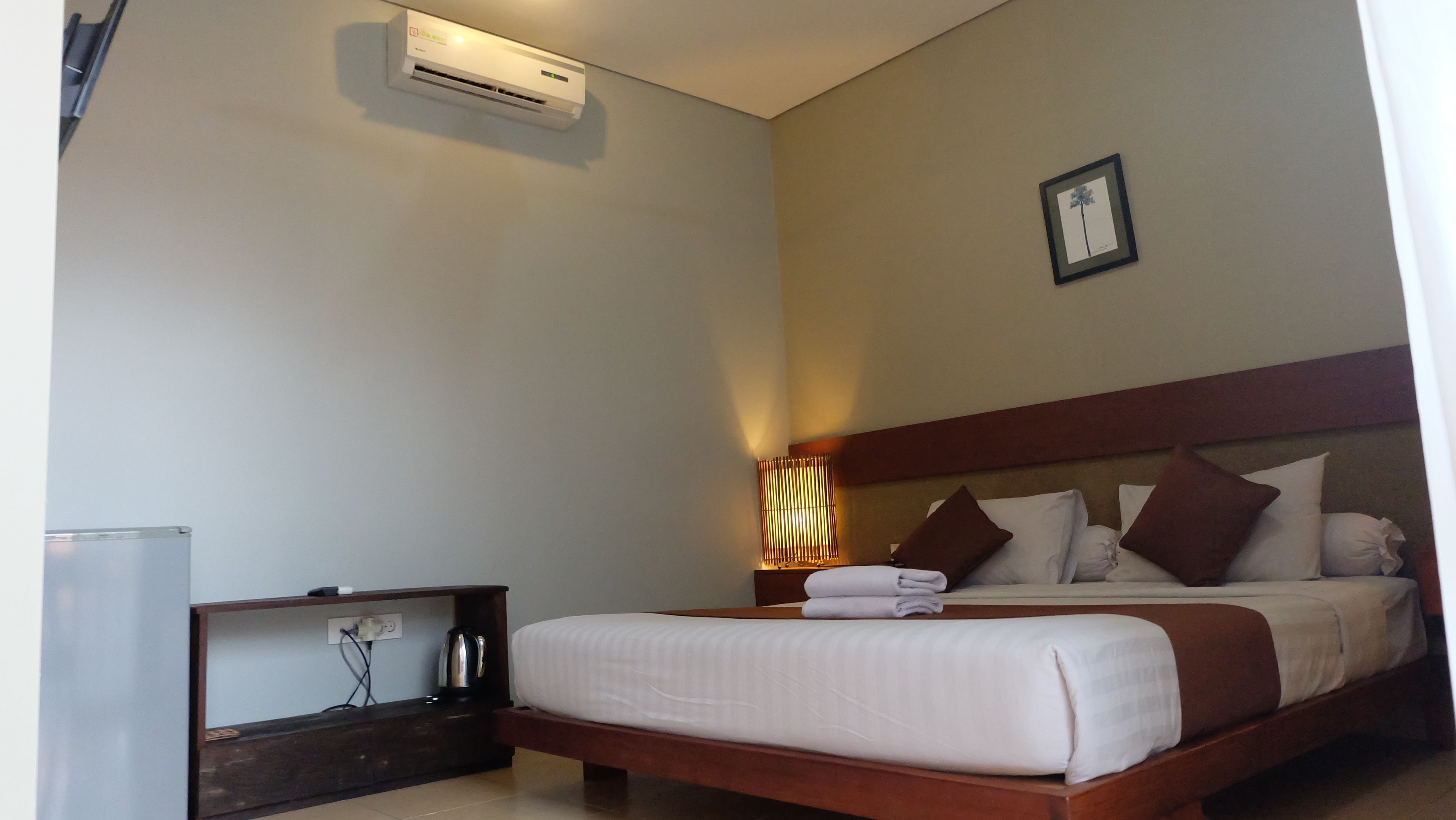 A hotel room with a bed, a bedside table with a kettle, an air conditioner, and a wall-mounted picture.