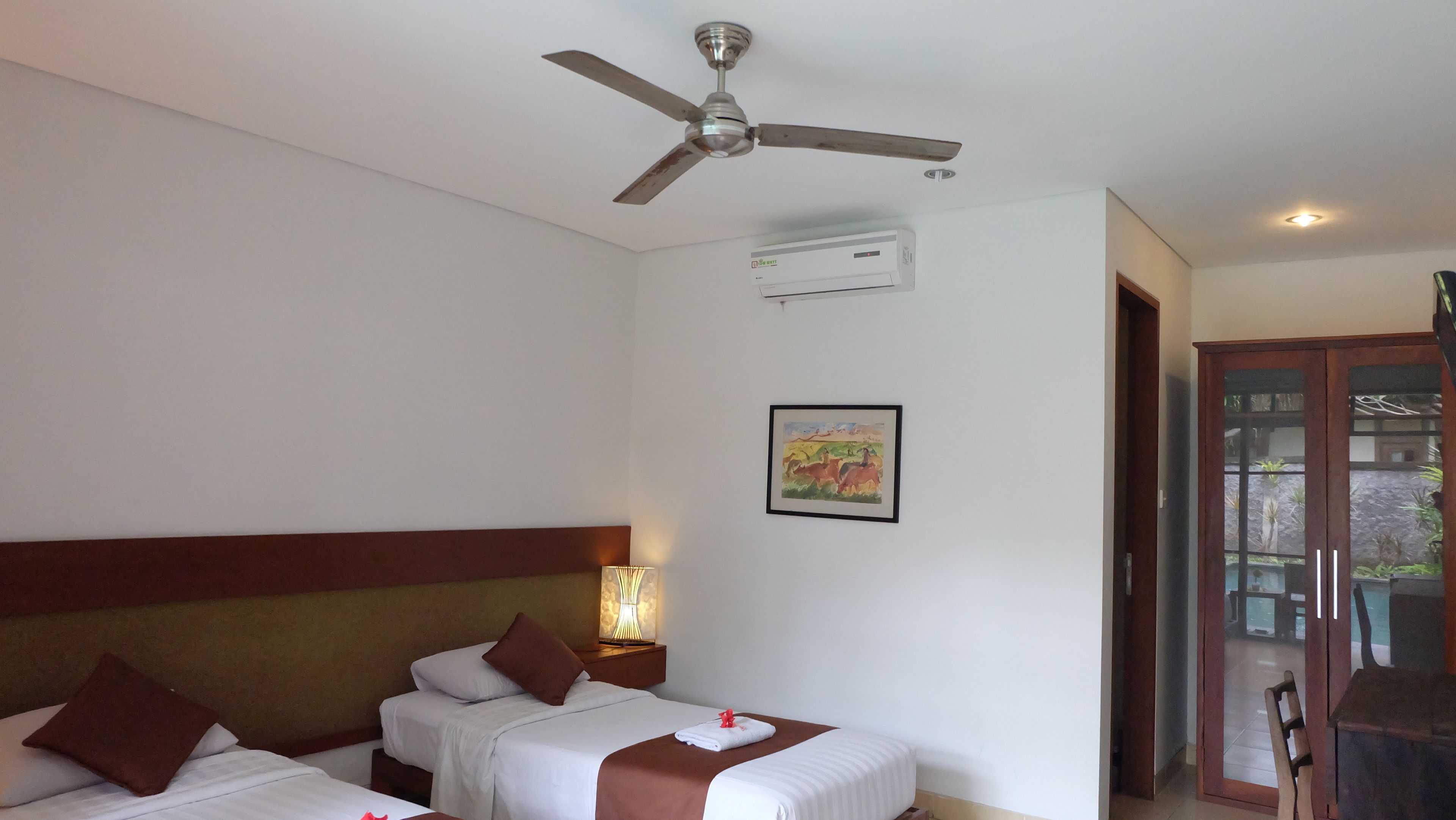 Quarto de hotel com duas camas, um ventilador de teto, ar condicionado, um quadro e vista para o exterior através de uma porta de vidro.