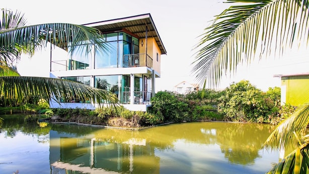 A modern two-story house with a glass facade, surrounded by lush greenery and a calm pond. - Plern Salaya Resort (Phutthamonthon)