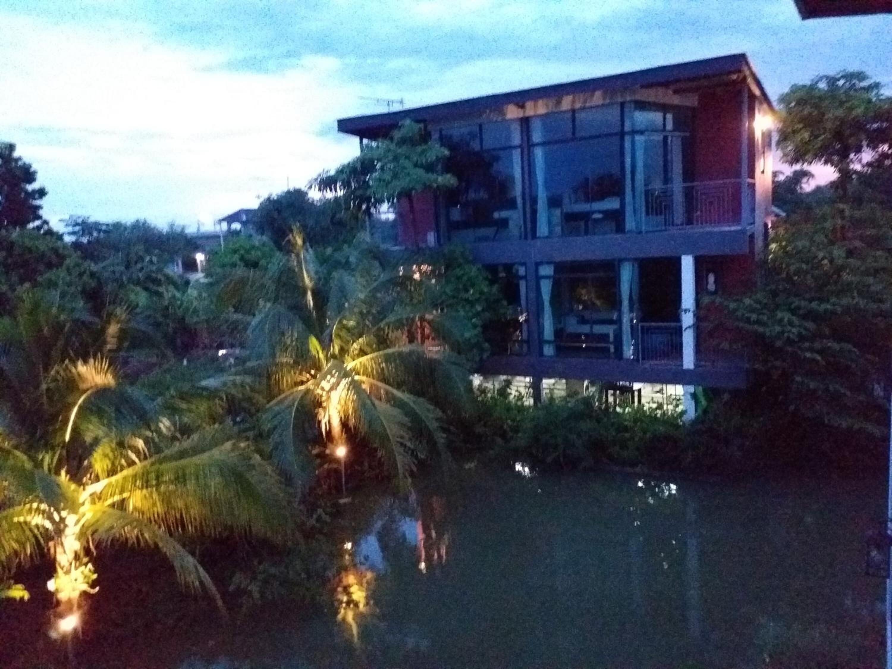 A modern two-story house with a balcony, surrounded by lush greenery and a body of water.
