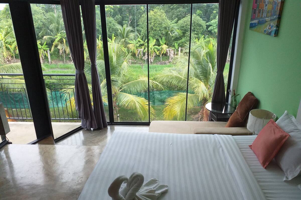 A hotel room with a large bed, a view of a lush garden through a sliding glass door, and a green wall with a painting.