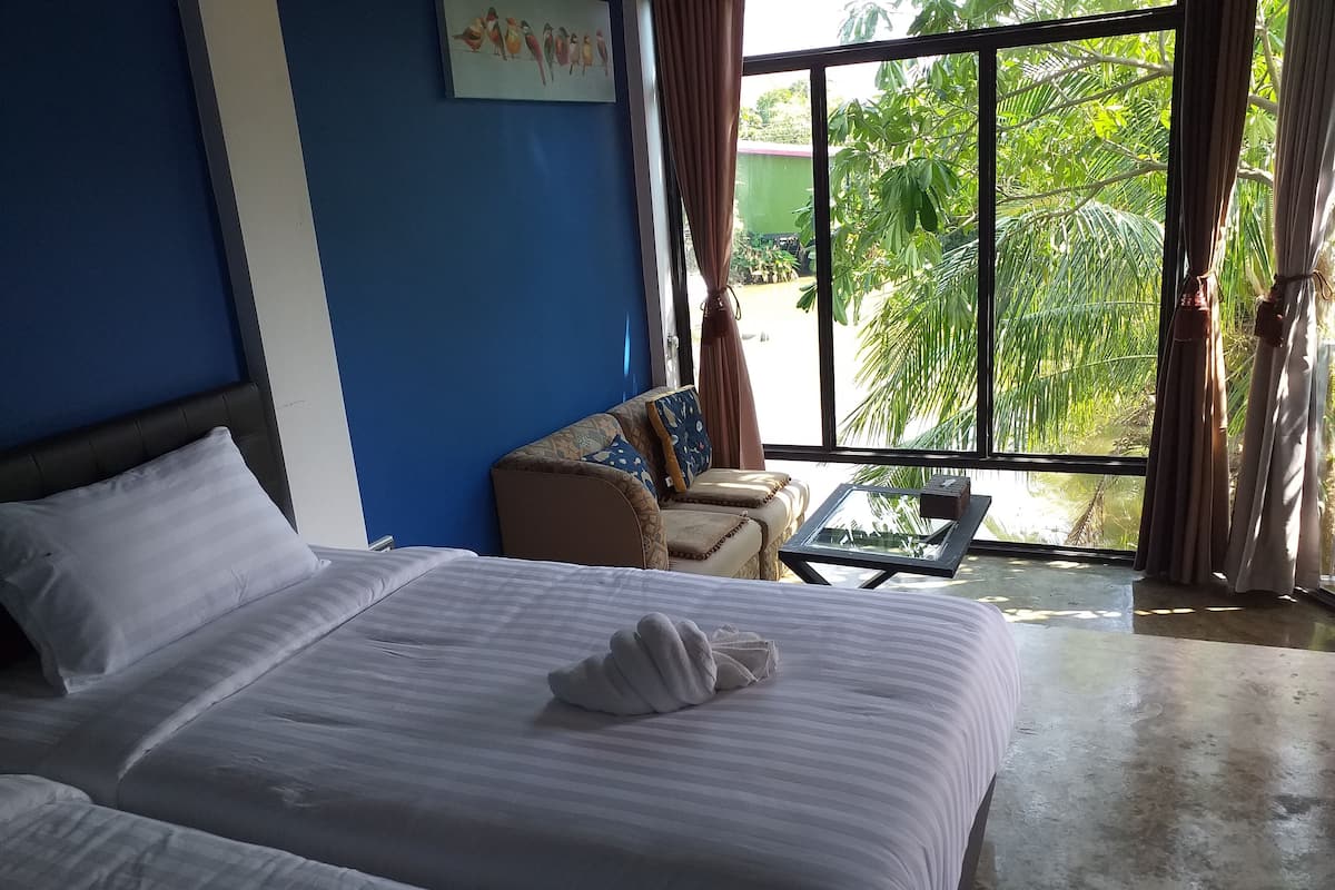 A hotel room with a large bed, a small sofa, a glass table, and a view of greenery outside.